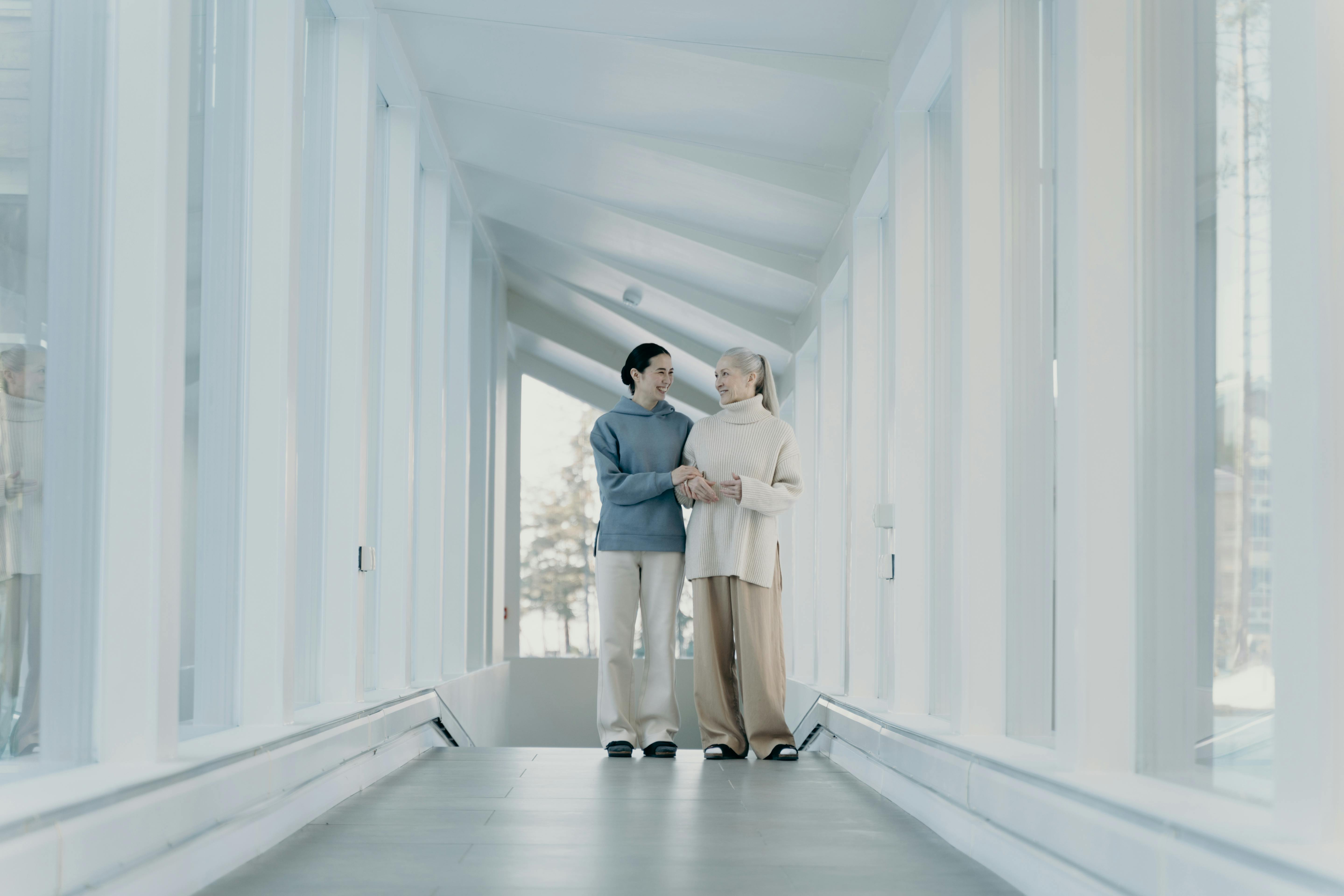 Caregiver walking arm-in-arm with elderly woman in a bright corridor